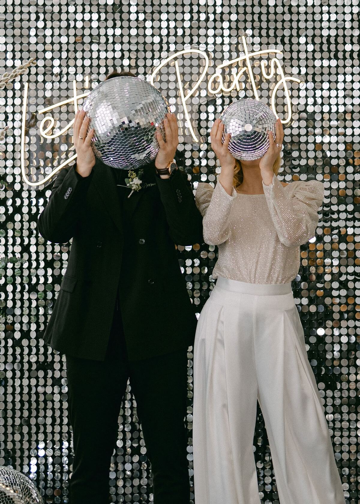 Bride and groom hold disco balls in front of their faces at a stylish wedding party with a glittering backdrop and a 'Let's Party' neon sign