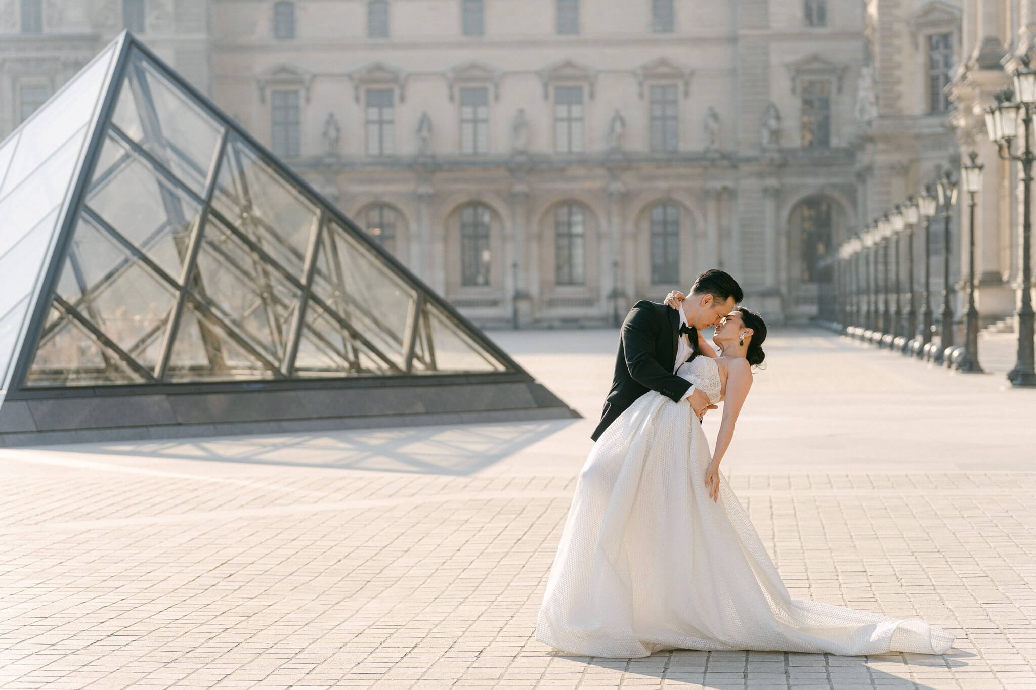 Bridal couple lovingly kissing in front of the Louvre Museum in Paris with the famous glass pyramid in the background at sunrise