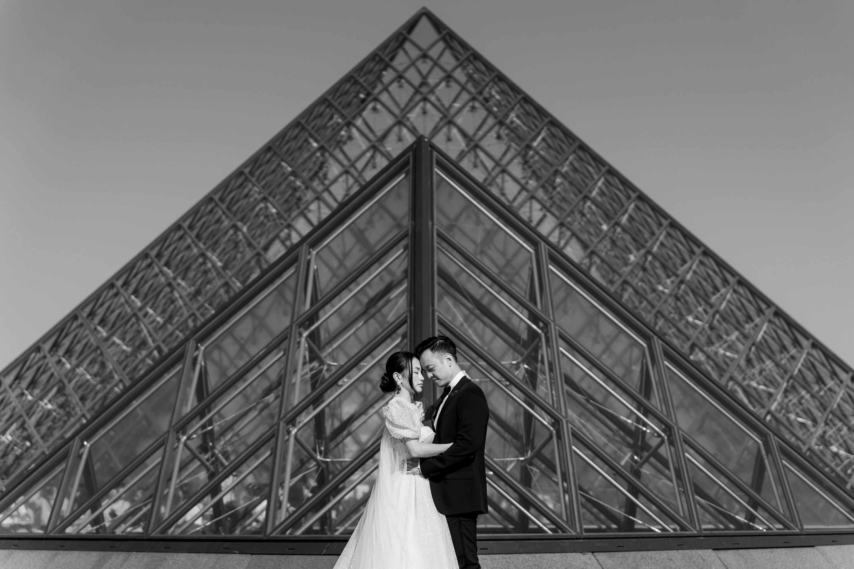 Schwarz-weißes Foto eines Brautpaares, das sich zärtlich vor der Glaspyramide des Louvre in Paris umarmt.
