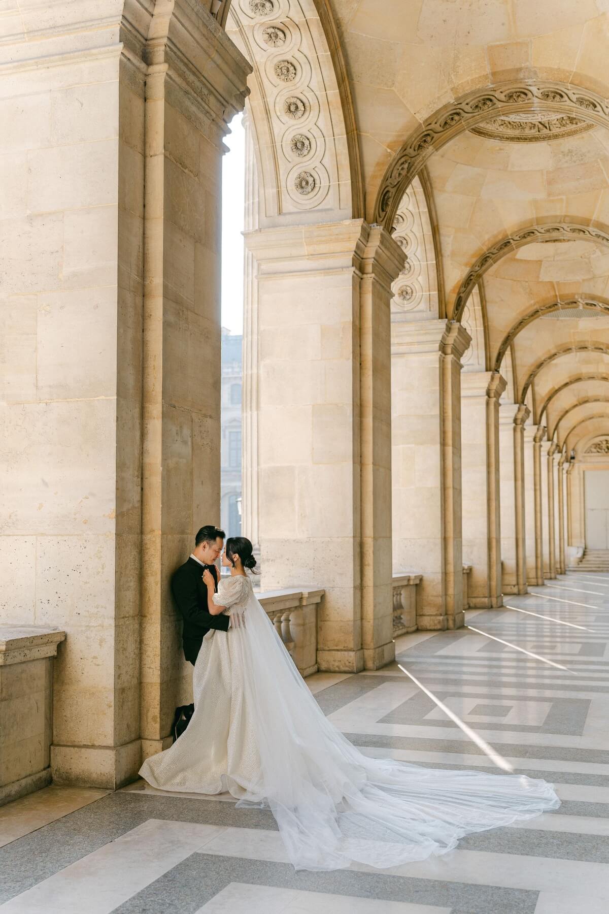 A bride and groom stand tenderly together in a romantic pose under the historic arches of the Louvre in Paris, surrounded by dramatic architecture and natural light.