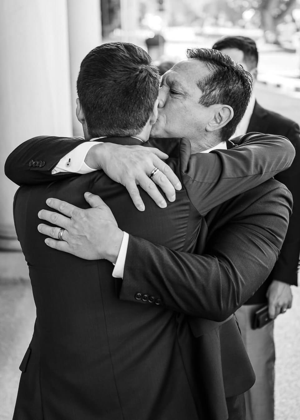 Emotional embrace and kiss on the forehead between father and son in elegant suits at a wedding celebration in the Kurhaus Wiesbaden in black and white.